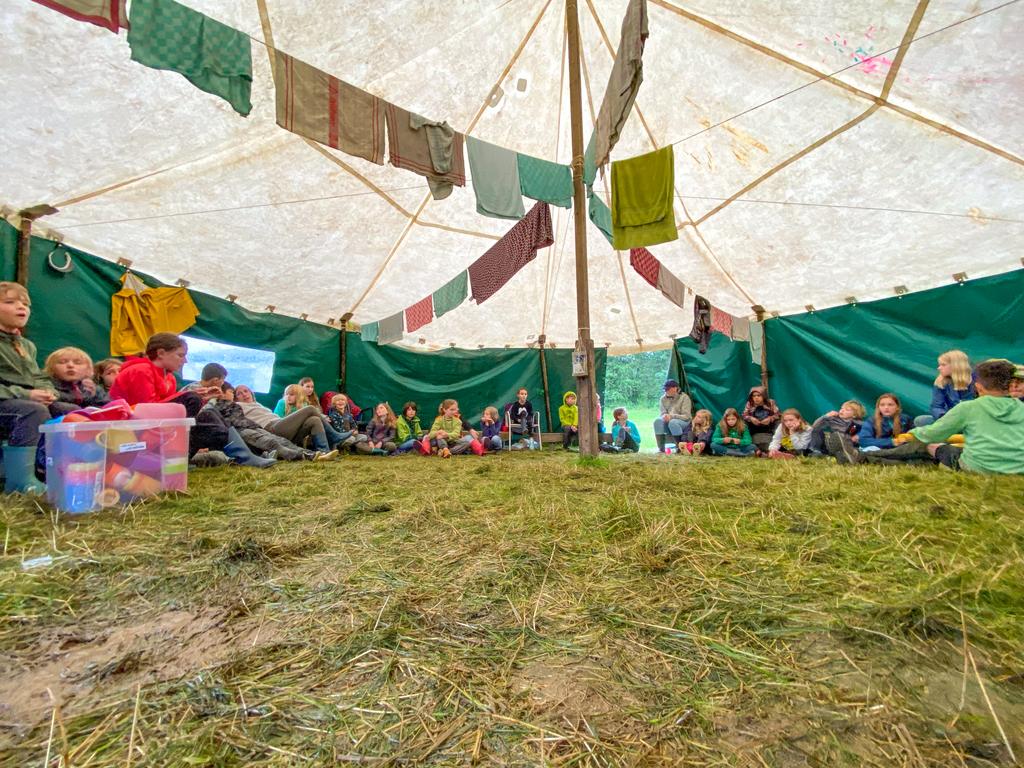Kinderen en leiding zitten in een cirkel in een grote tent waar keukenhandoeken als slingers hangen te drogen.