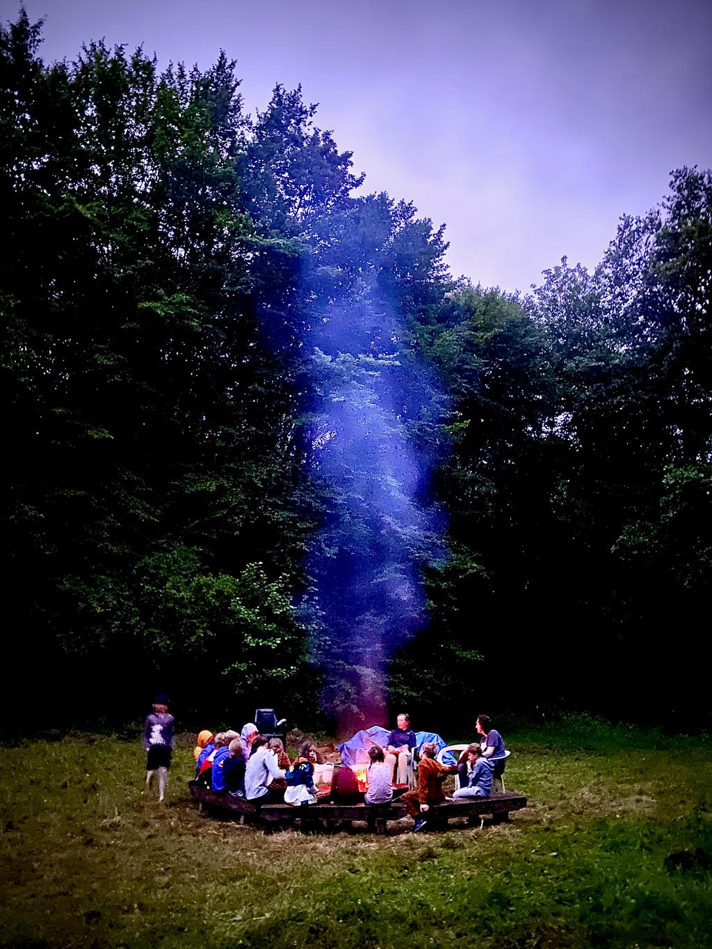 Een groep leiding en snuisters zitten knus rond een kampvuur waarvan de rook zichtbaar is tegen een achtergrond van bomen.