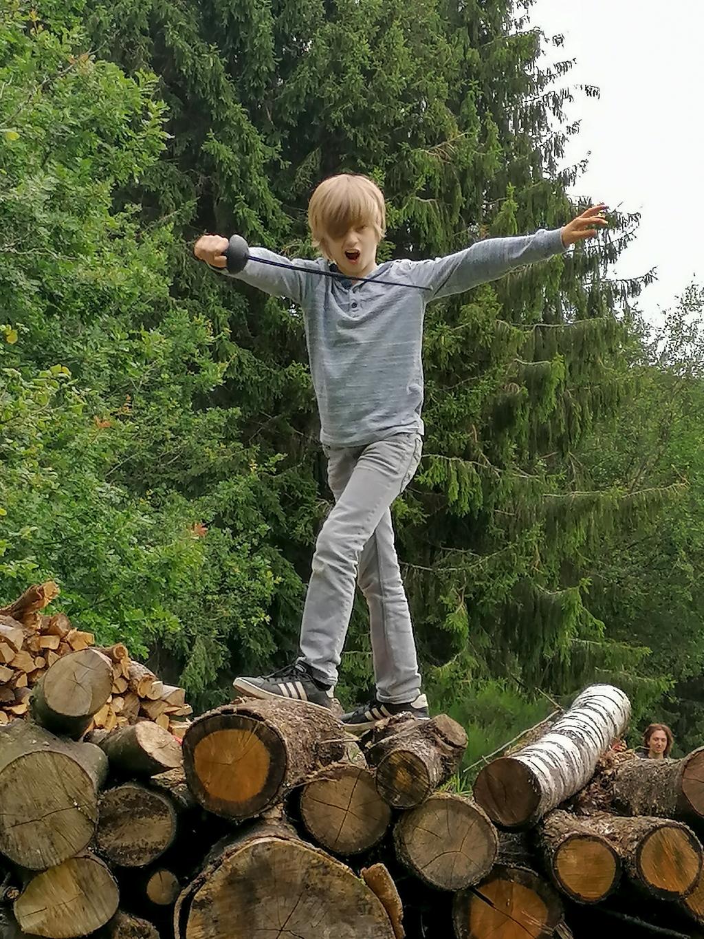 Een blond kind staat op een stapel hout met een getrokken plastieken sabel.