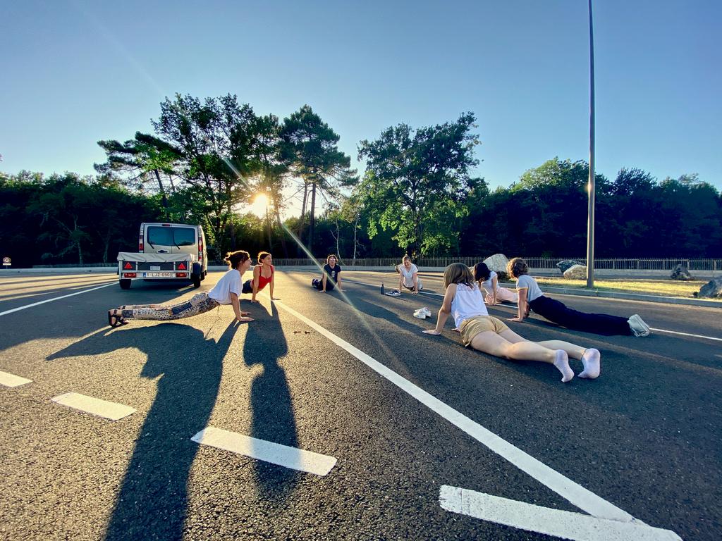 Zeven mensen houden in een cirkel een yoga-pose aan op een snelwegparking terwijl de avondzon door de bomen heen straalt.